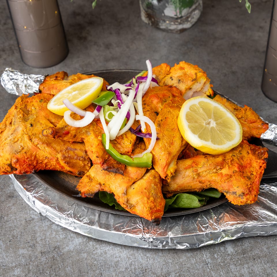 Tandoori chicken pieces with lemon, onions, and peppers on a dark plate with foil.