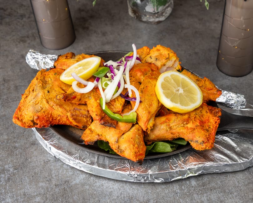 Tandoori chicken pieces with lemon, onions, and peppers on a dark plate with foil.
