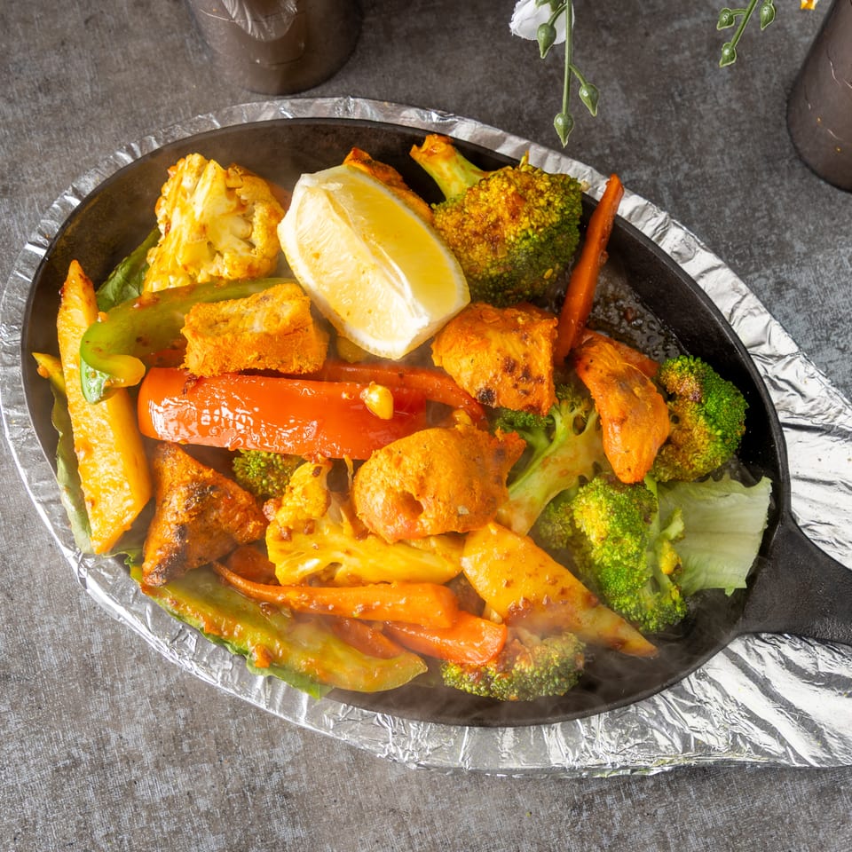 Sizzling platter of chicken tikka, broccoli, cauliflower, carrots, and bell peppers with a lemon wedge.