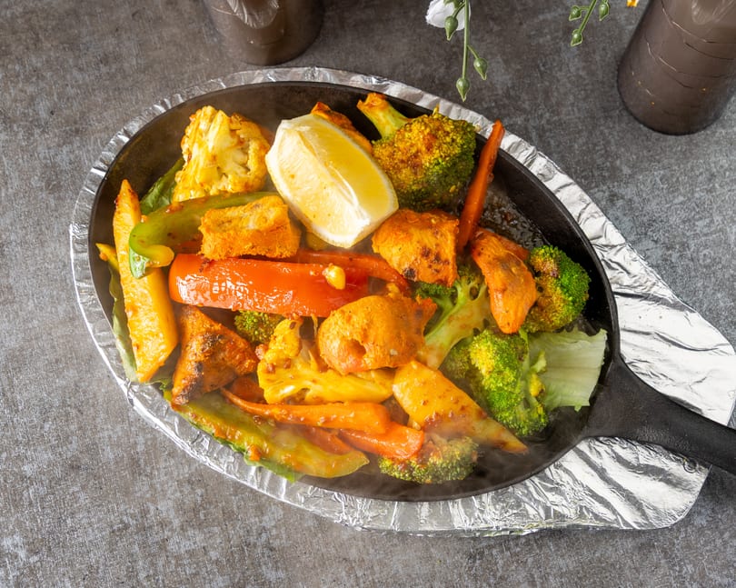 Sizzling platter of chicken tikka, broccoli, cauliflower, carrots, and bell peppers with a lemon wedge.