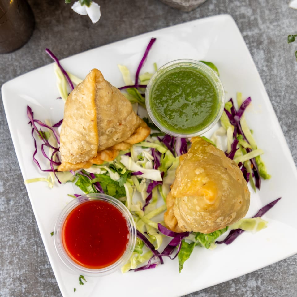 Two golden-brown samosas on a white plate with shredded cabbage, green and red dipping sauces.