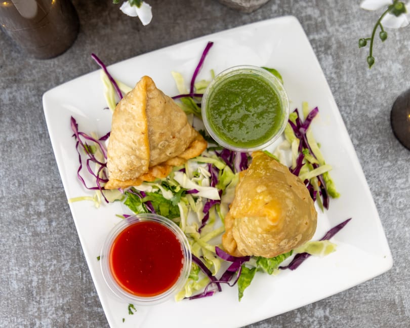 Two golden-brown samosas on a white plate with shredded cabbage, green and red dipping sauces.