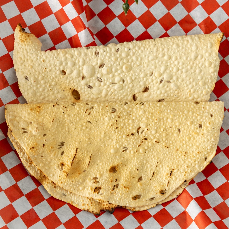 Two crispy papadum flatbreads with cumin seeds, served on red and white checkered paper.
