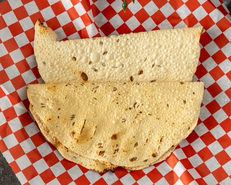 Two crispy papadum flatbreads with cumin seeds, served on red and white checkered paper.