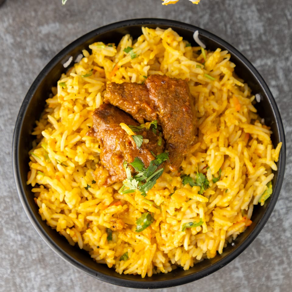 Overhead view of a black bowl filled with yellow biryani rice and three pieces of meat.