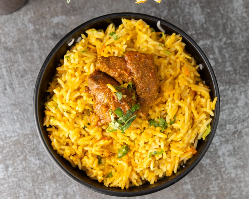Overhead view of a black bowl filled with yellow biryani rice and three pieces of meat.