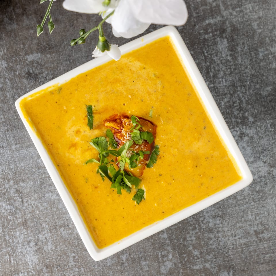 Square white bowl of yellow soup garnished with herbs and a crouton.