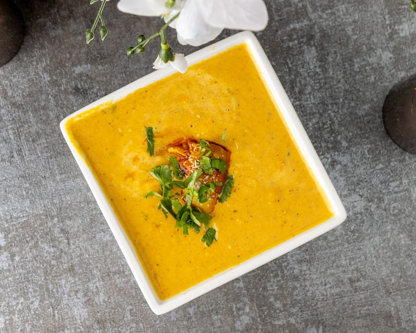 Square white bowl of yellow soup garnished with herbs and a crouton.