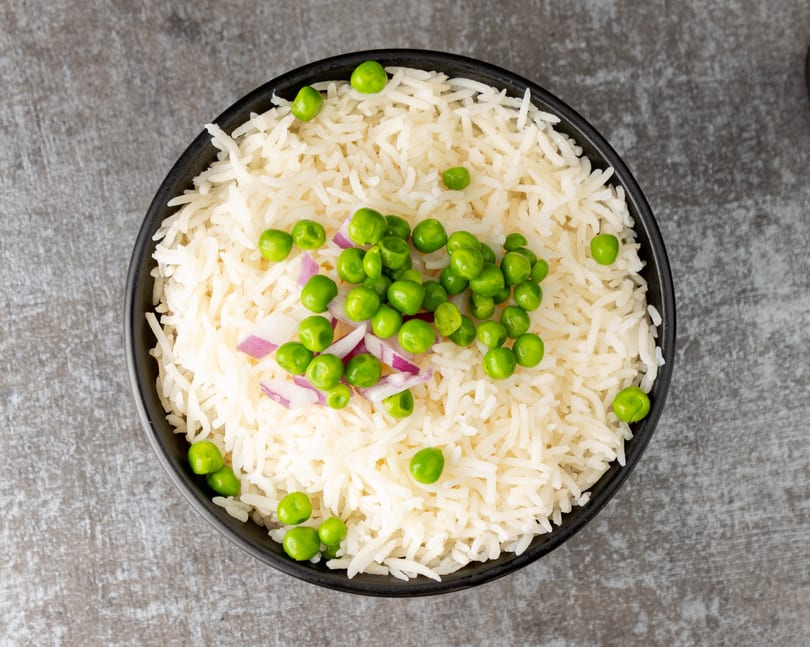 Overhead view of a black bowl filled with white rice, green peas, and red onion.