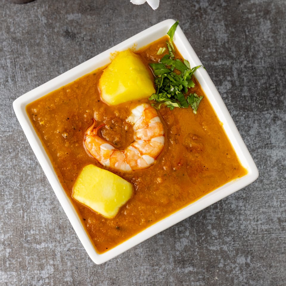 A white square bowl filled with a rich orange curry, shrimp, potatoes, and cilantro.