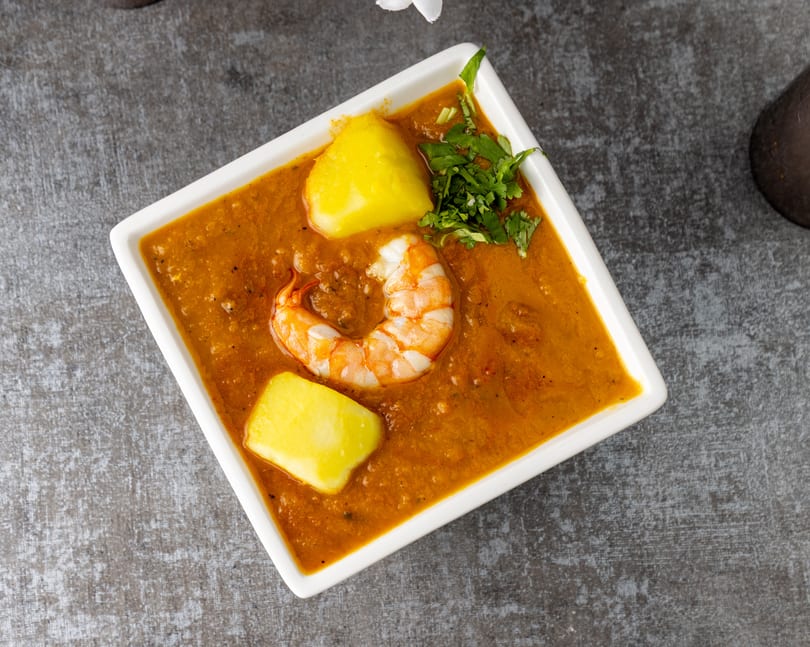 A white square bowl filled with a rich orange curry, shrimp, potatoes, and cilantro.