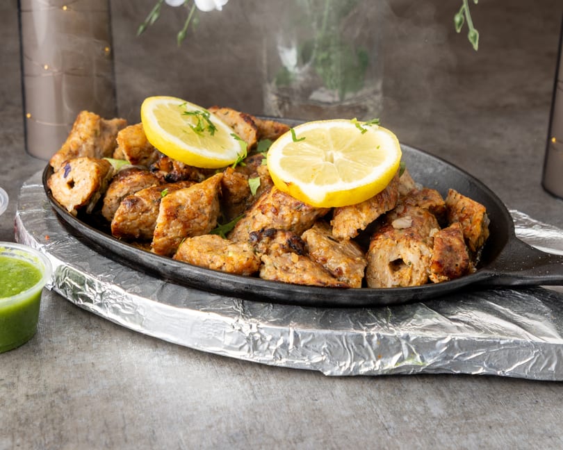 Sizzling chicken tikka with lemon slices and cilantro on a black platter, with green chutney.