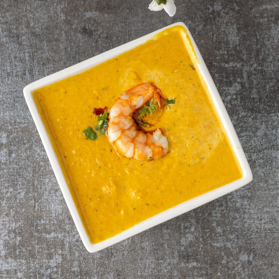 A white square bowl filled with vibrant yellow shrimp bisque, garnished with a single shrimp and herbs.