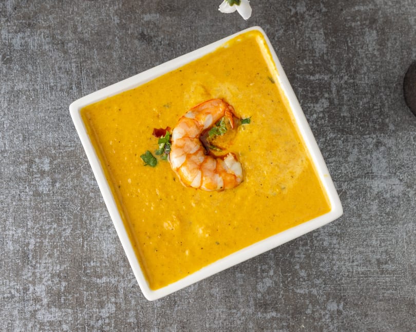 A white square bowl filled with vibrant yellow shrimp bisque, garnished with a single shrimp and herbs.