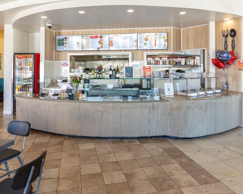 Restaurant interior with a long counter, menu boards, a Coca-Cola fridge, and "Happy Birthday" balloons.