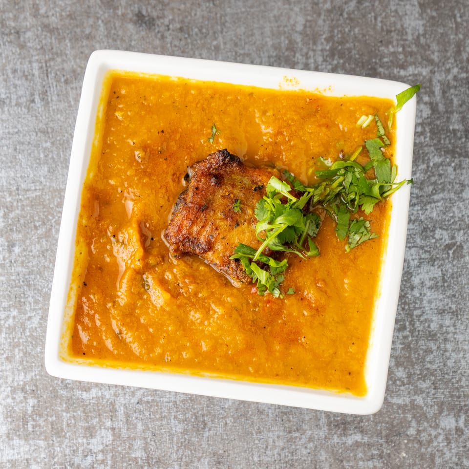 A white rectangular bowl filled with orange curry, topped with a piece of meat and fresh cilantro.