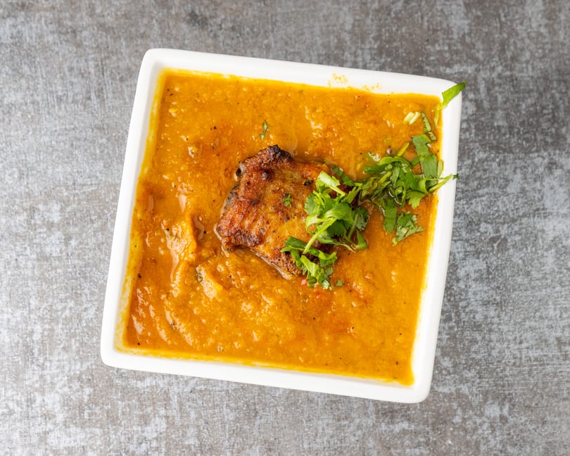 A white rectangular bowl filled with orange curry, topped with a piece of meat and fresh cilantro.