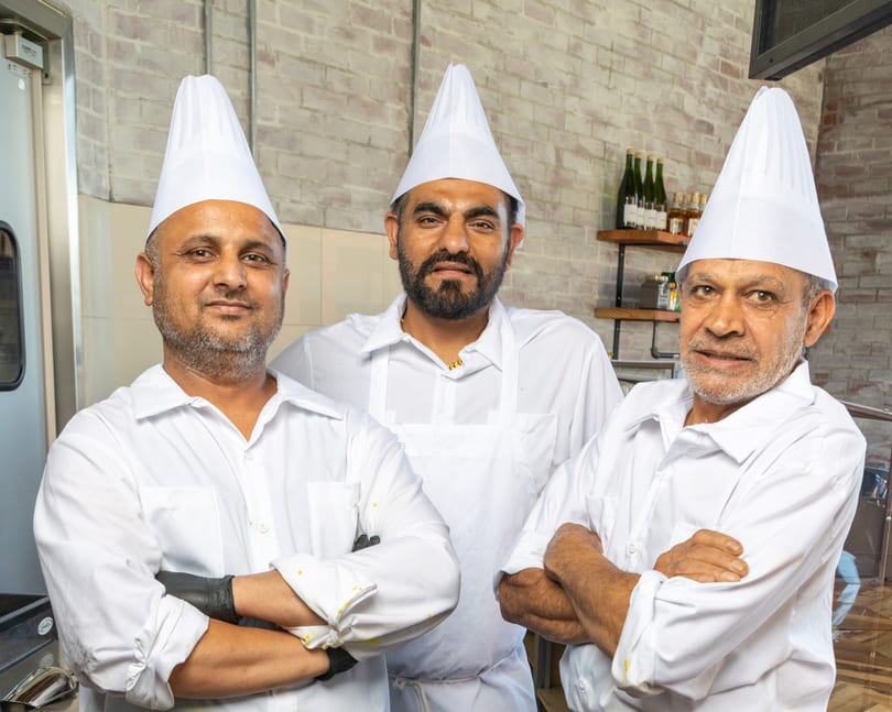 Three male chefs in white uniforms and hats stand with arms crossed in a kitchen.