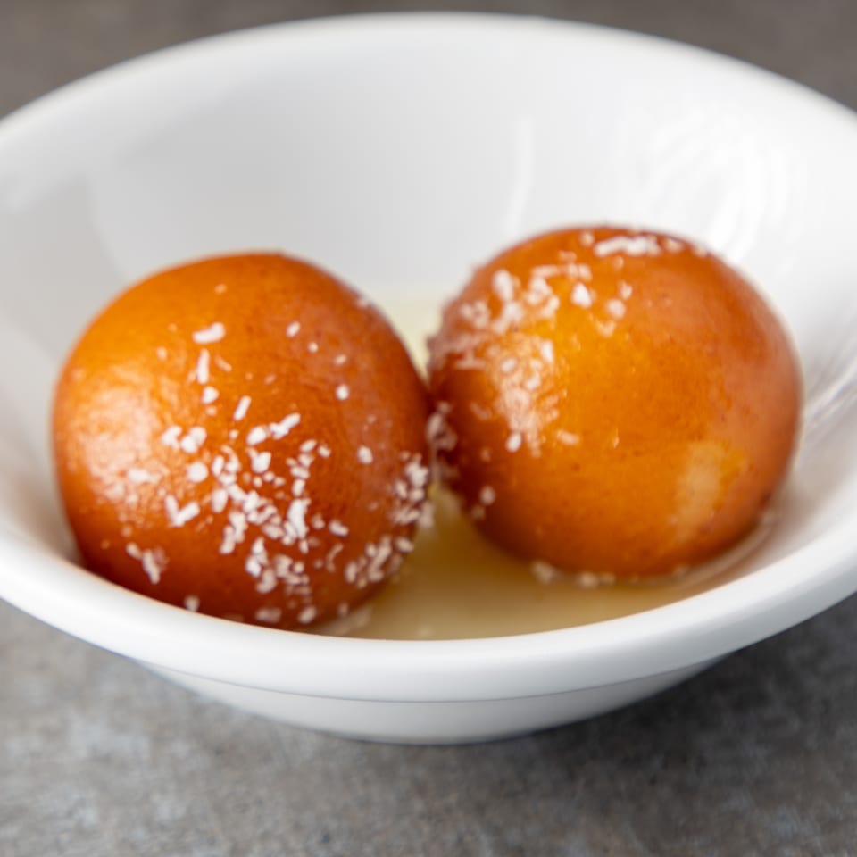 Two gulab jamun, garnished with coconut flakes, sit in a white bowl with syrup.