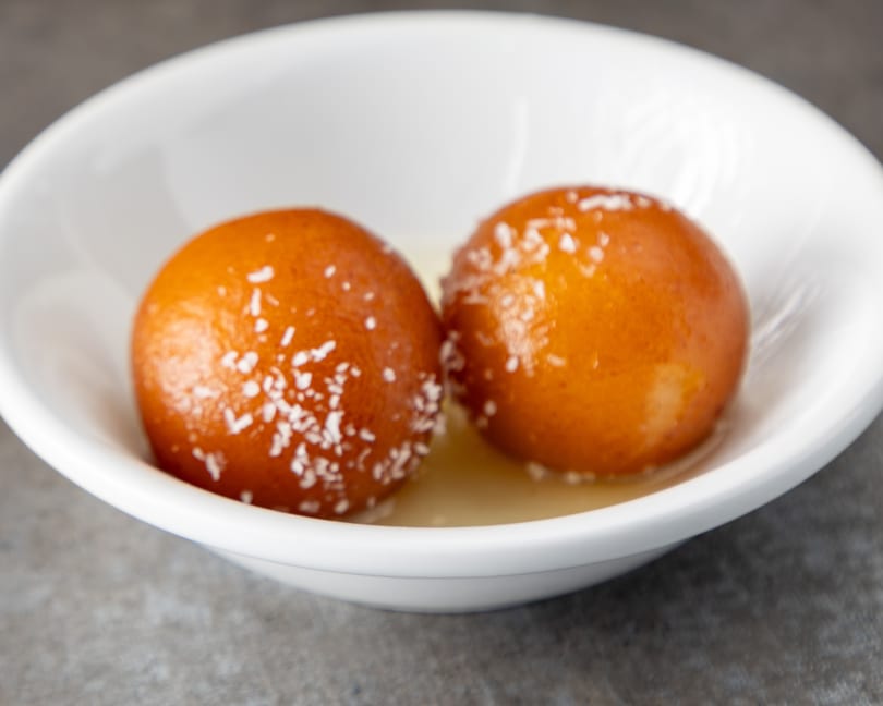 Two gulab jamun, garnished with coconut flakes, sit in a white bowl with syrup.