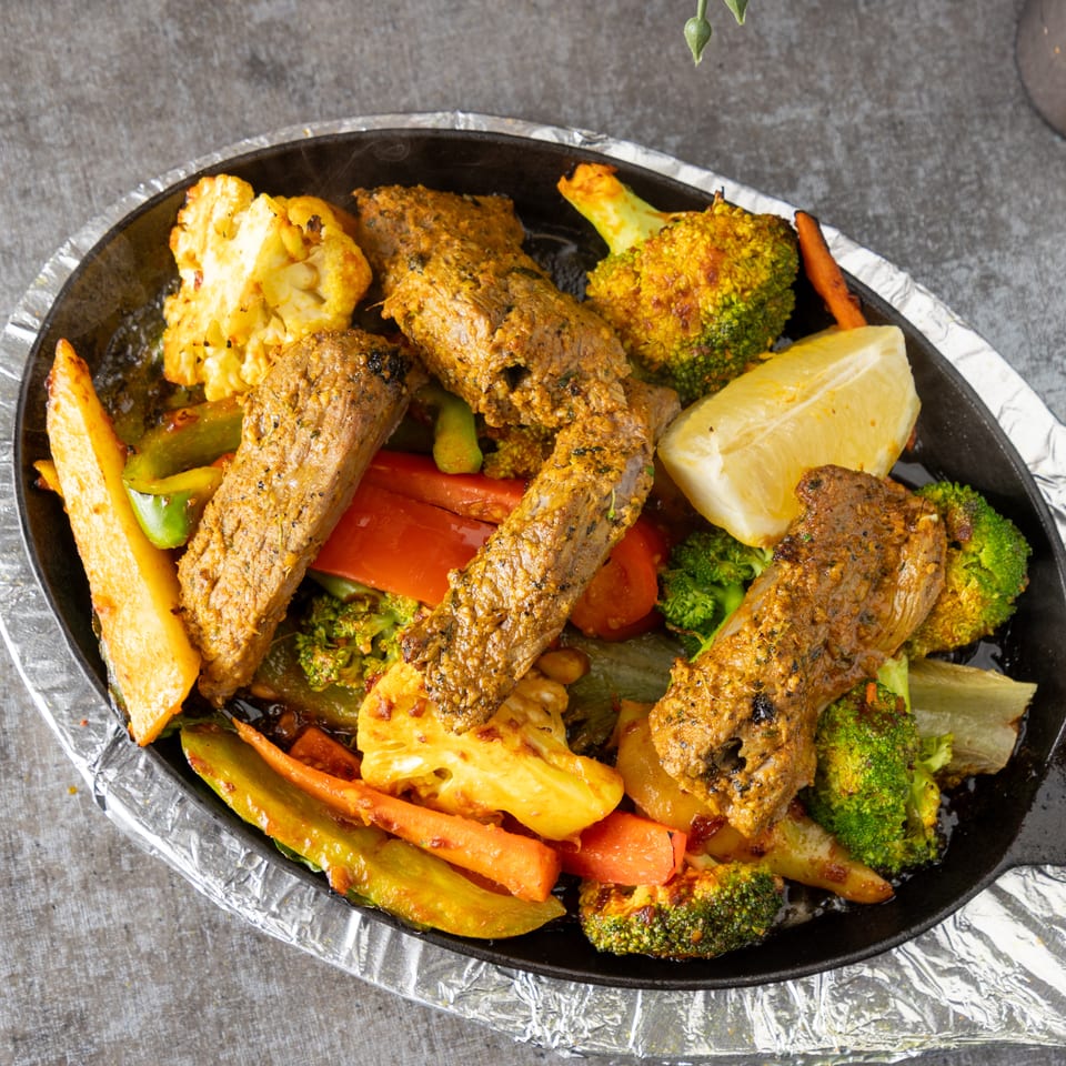 Sizzling platter of seasoned meat strips, broccoli, cauliflower, carrots, and bell peppers with a lemon wedge.