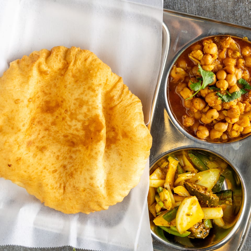 A golden fried puri bread, chana masala, and mixed vegetable pickle on a metal tray.