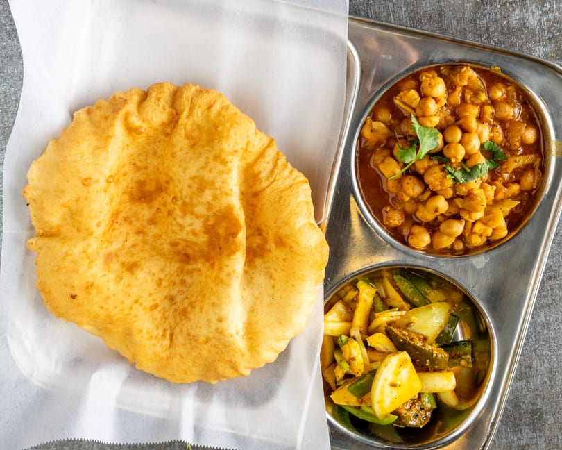 A golden fried puri bread, chana masala, and mixed vegetable pickle on a metal tray.