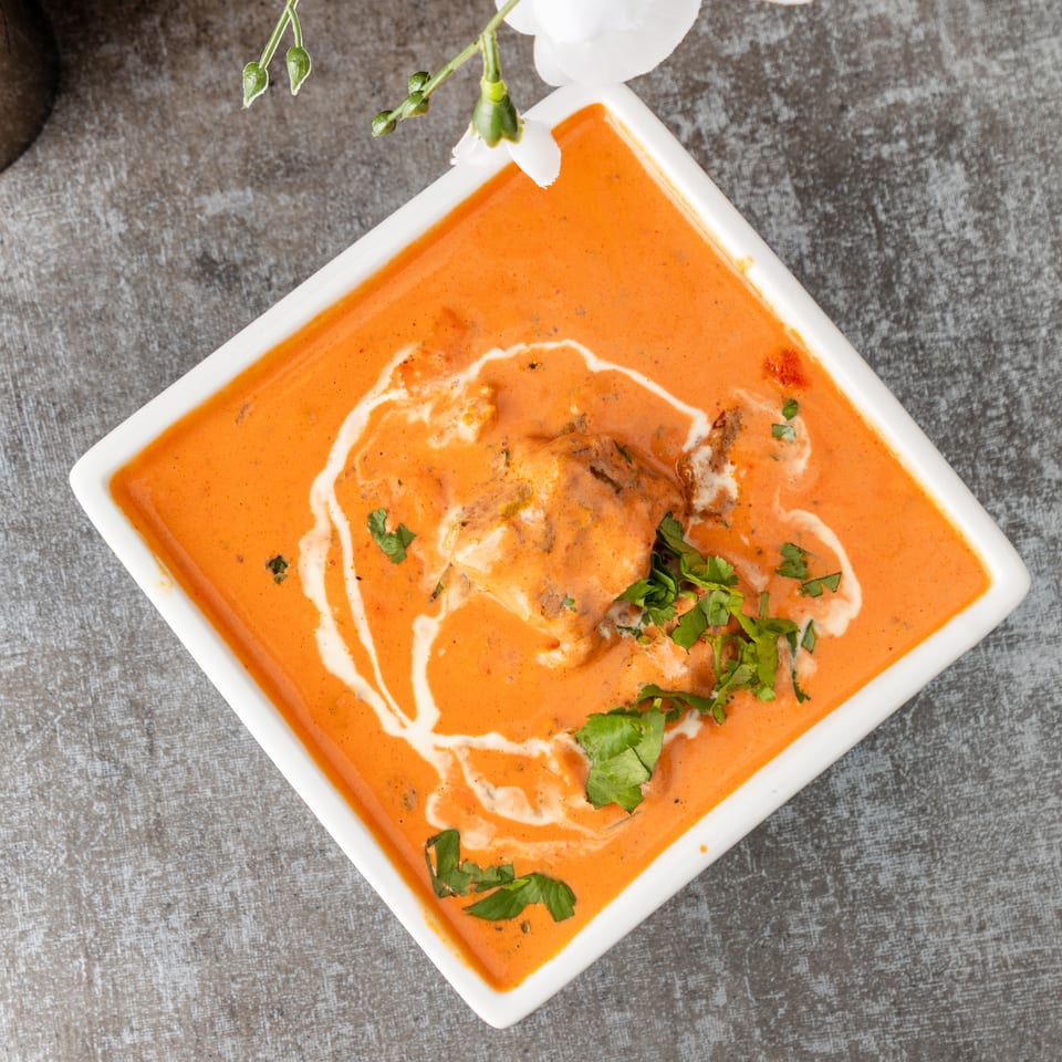 Overhead view of butter chicken in a white square bowl, garnished with cream swirls and fresh cilantro.