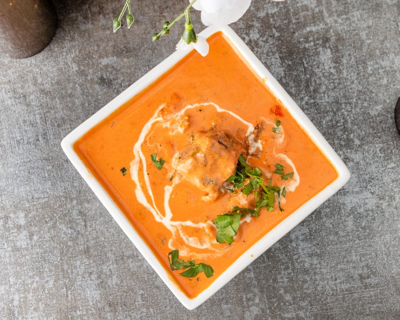 Overhead view of butter chicken in a white square bowl, garnished with cream swirls and fresh cilantro.