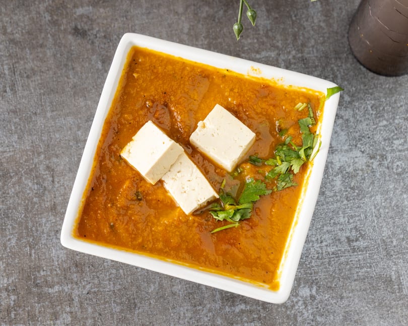 Overhead view of a white rectangular dish filled with orange curry, topped with paneer and cilantro.