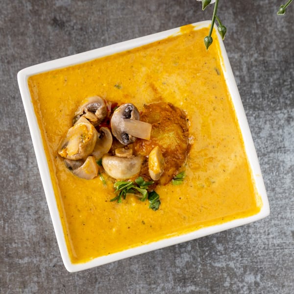 Creamy orange soup with sautéed mushrooms, a golden fritter, and fresh herbs in a white bowl.