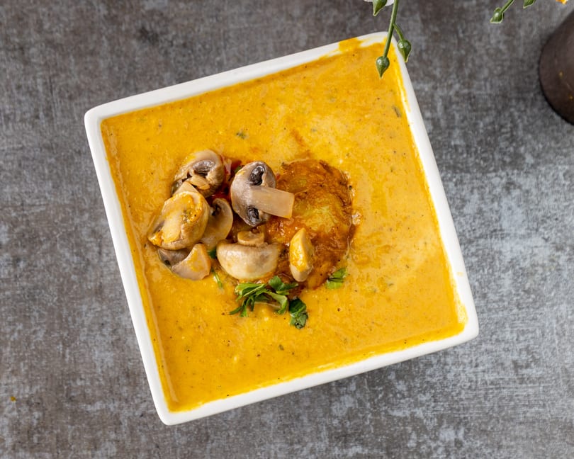 Creamy orange soup with sautéed mushrooms, a golden fritter, and fresh herbs in a white bowl.