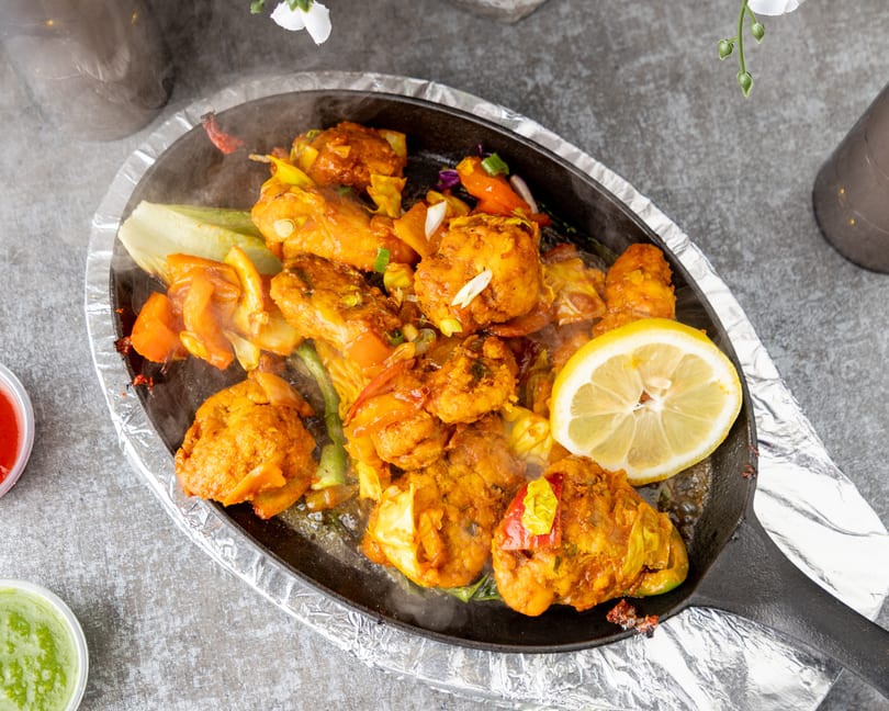 Sizzling chicken tikka with vegetables and a lemon slice on a hot plate.