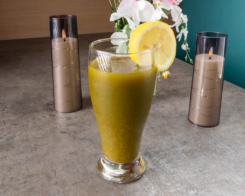 Green smoothie with a lemon slice, flanked by two decorative candles and flowers on a table.