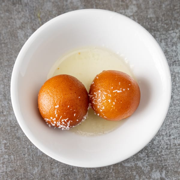 Two gulab jamun, Indian fried dough balls, in a white bowl with syrup and coconut flakes.