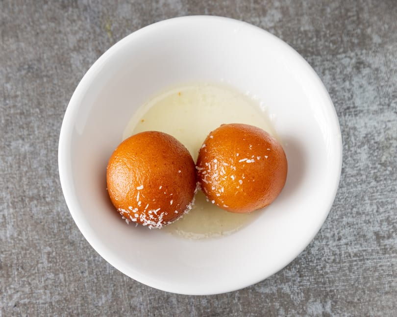 Two gulab jamun, Indian fried dough balls, in a white bowl with syrup and coconut flakes.