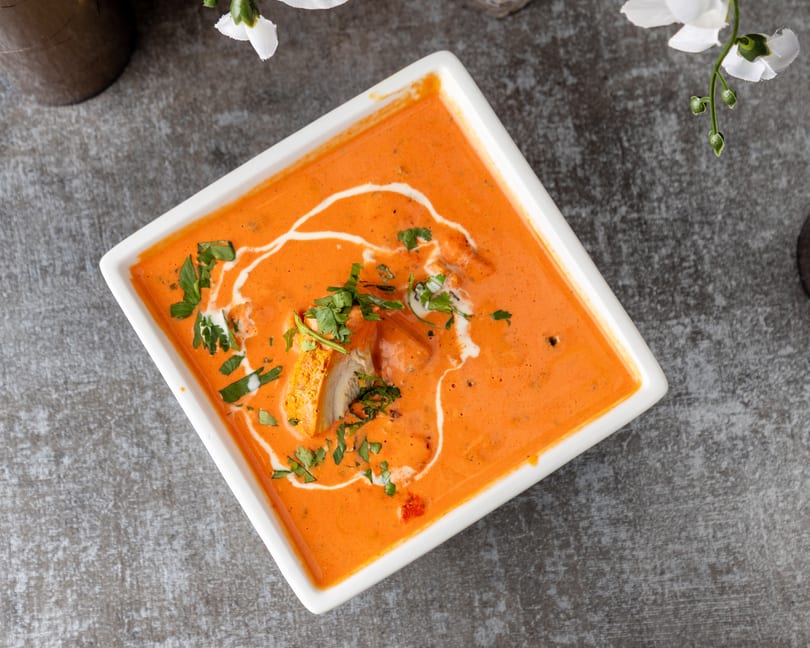 Overhead view of butter chicken in a white square bowl, garnished with cream swirls and cilantro.