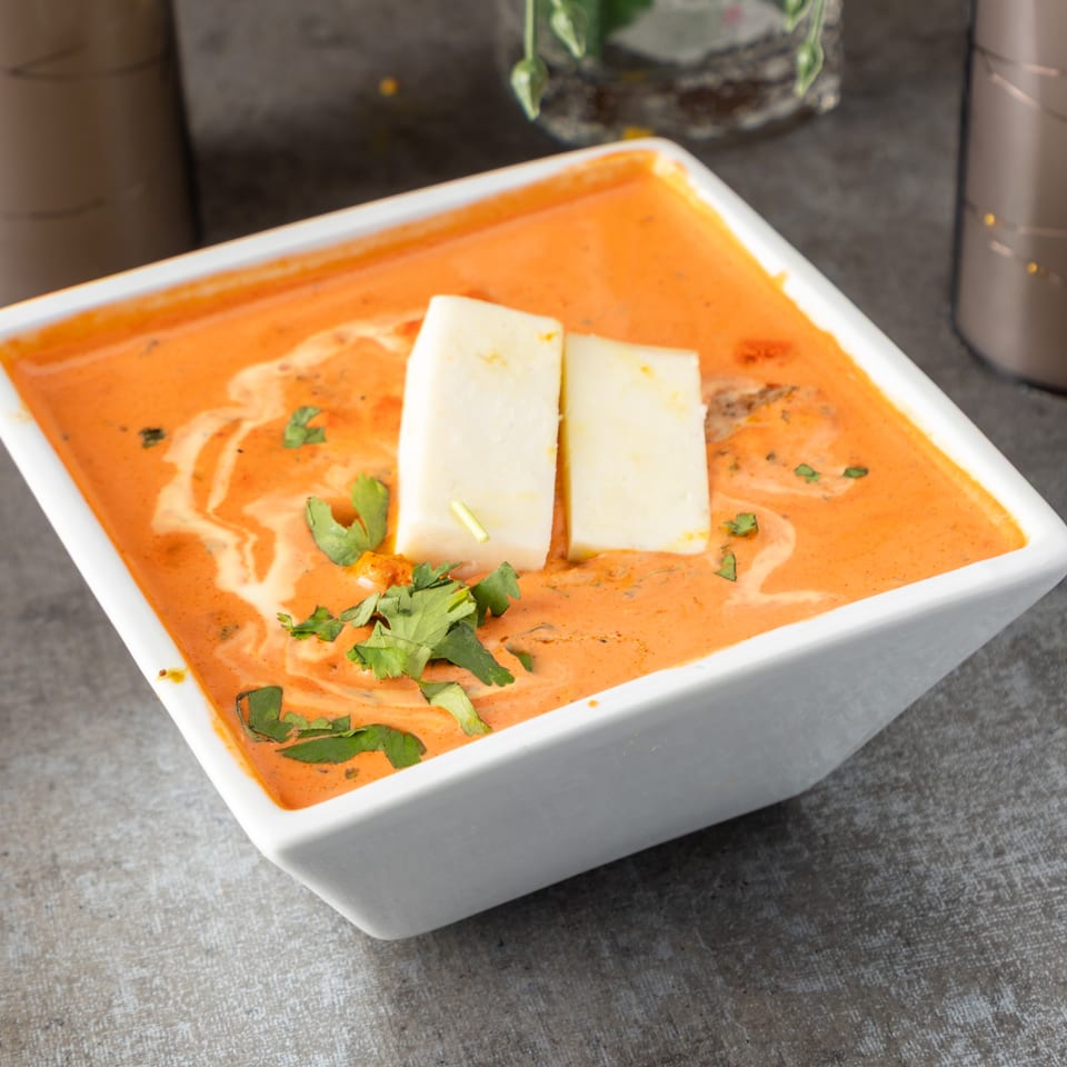 A white square bowl filled with orange curry, topped with two paneer slices and fresh cilantro.