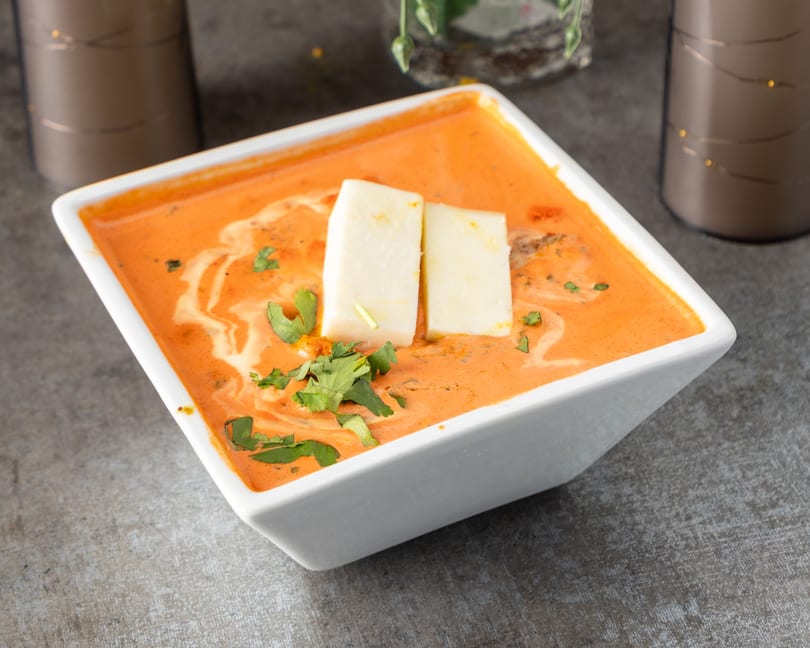 A white square bowl filled with orange curry, topped with two paneer slices and fresh cilantro.