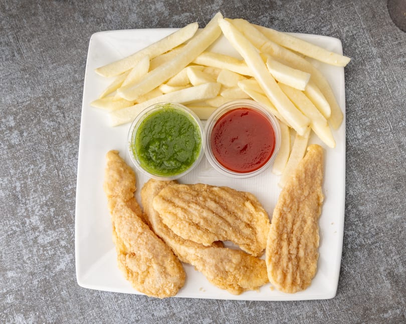 A white plate holds crispy chicken tenders, golden french fries, green chutney, and red ketchup.