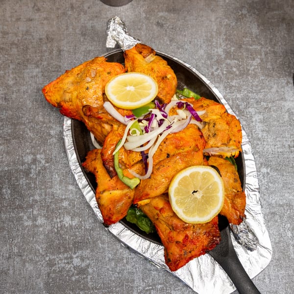 Sizzling tandoori chicken pieces with lemon, onions, and peppers on a black platter.