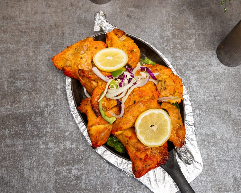 Sizzling tandoori chicken pieces with lemon, onions, and peppers on a black platter.